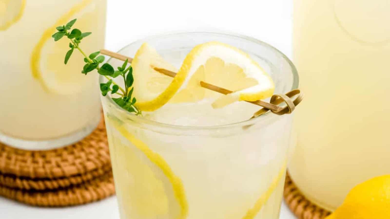 Glass of lemonade with ice, lemon slices, and a fresh herb garnish on a coaster, with more lemonade in the background.