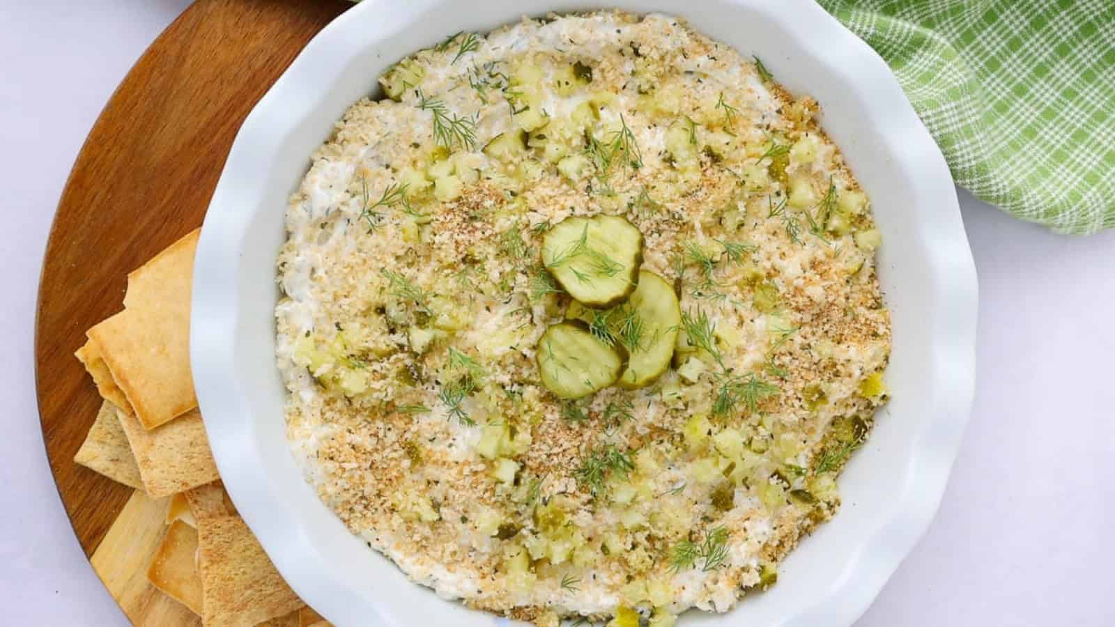 A baked dip topped with breadcrumbs, pickles, and dill, served with pita chips on a wooden tray.