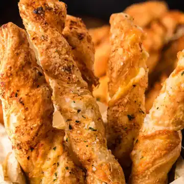 Close-up of golden, seasoned breadsticks twisted and served upright in a basket with dipping sauce nearby.