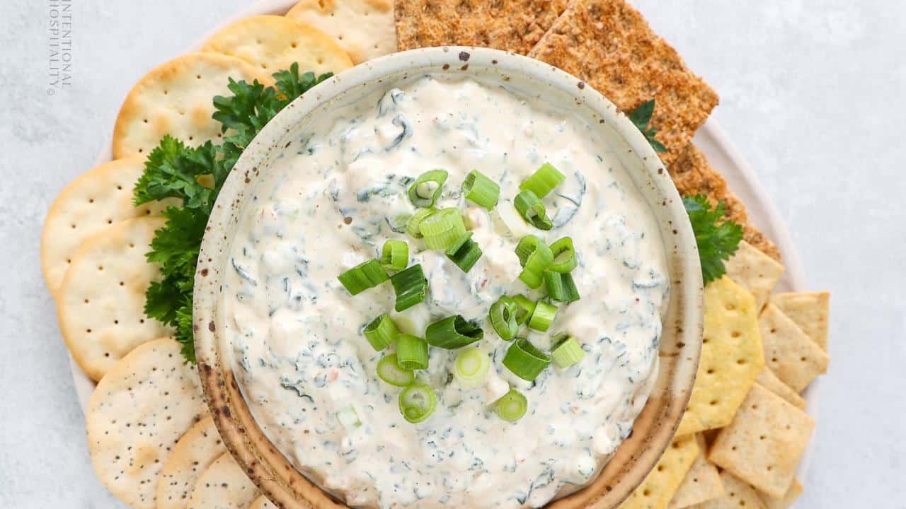 A bowl of creamy dip topped with chopped green onions, surrounded by assorted crackers and parsley.