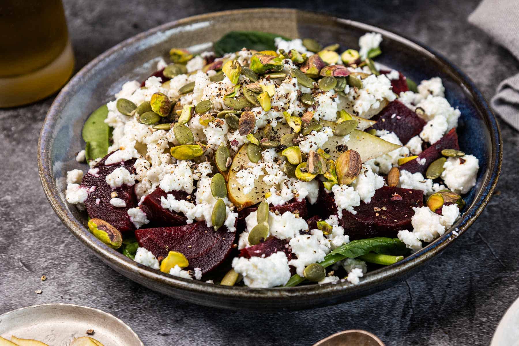 A bowl of beet salad with crumbled cheese, pears, spinach, pistachios, and pumpkin seeds.