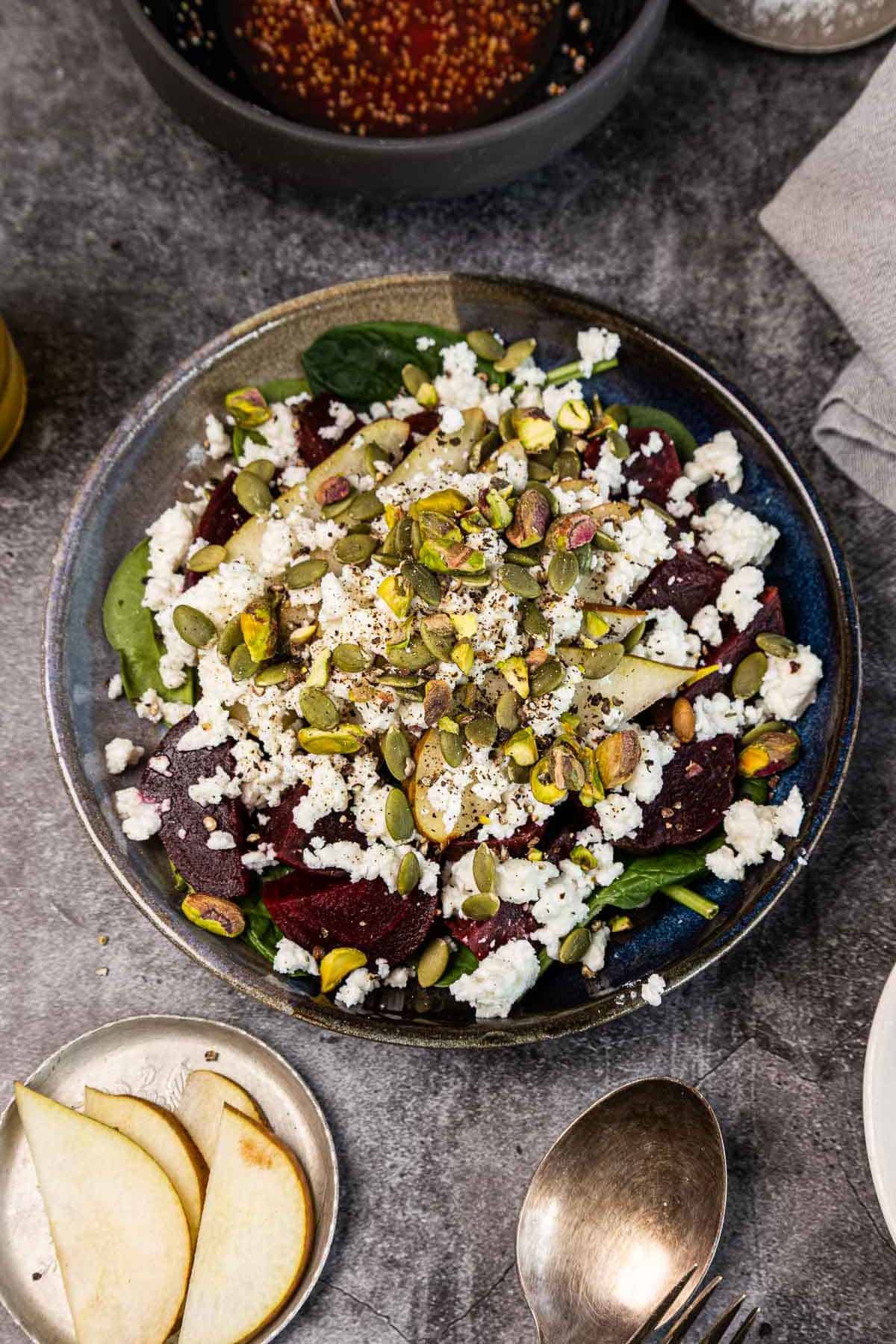 A salad with spinach, beets, feta cheese, pear slices, and pumpkin seeds in a dark bowl on a gray surface.