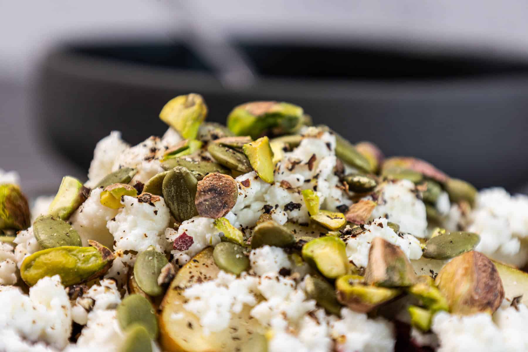 Close-up of a salad topped with crumbled cheese, pistachios, and pumpkin seeds, with a blurred bowl in the background.