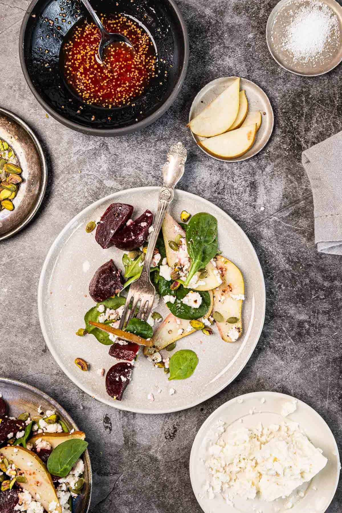 Overhead view of a beet, spinach, and pear salad with feta and pistachios on a gray table with dressing and ingredients.