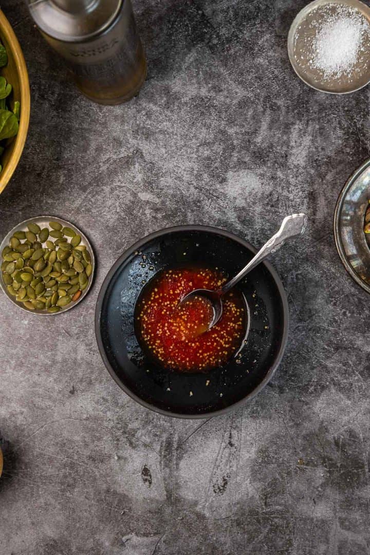 A bowl of red chili vinaigrette with a spoon, surrounded by pepitas and salt on a gray surface.