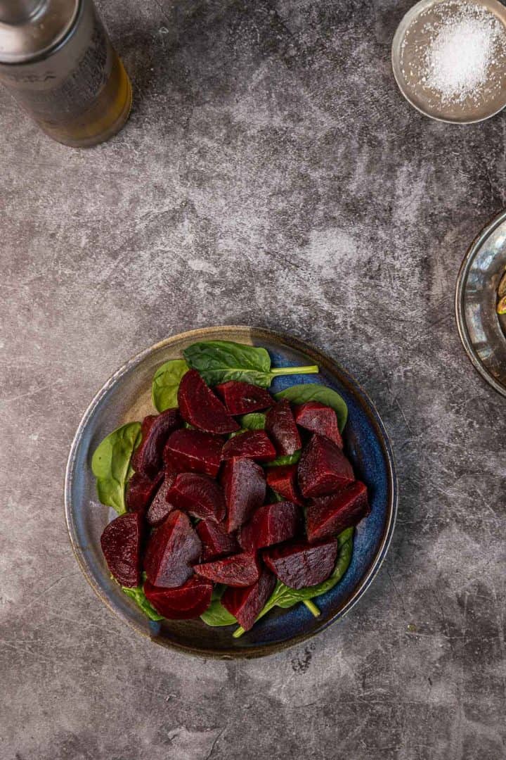 A plate of sliced beets on a bed of spinach, with olive oil and salt nearby on a gray surface.