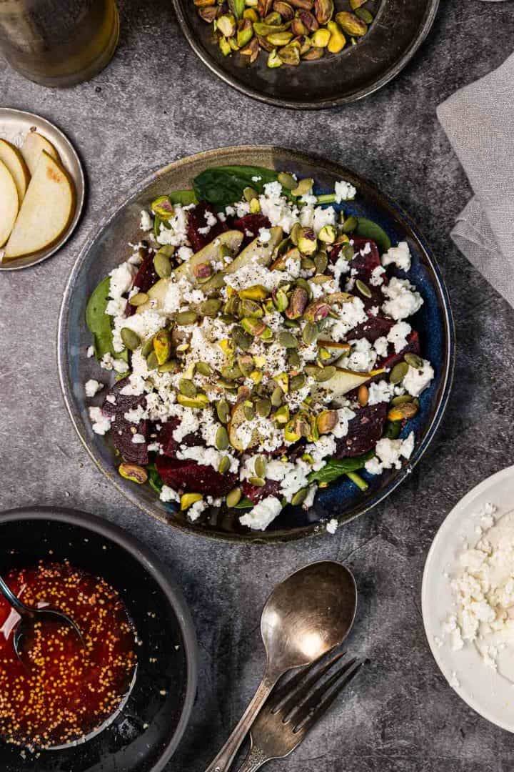A salad with beets, apple slices, feta cheese, and pistachios on a plate, surrounded by ingredients.