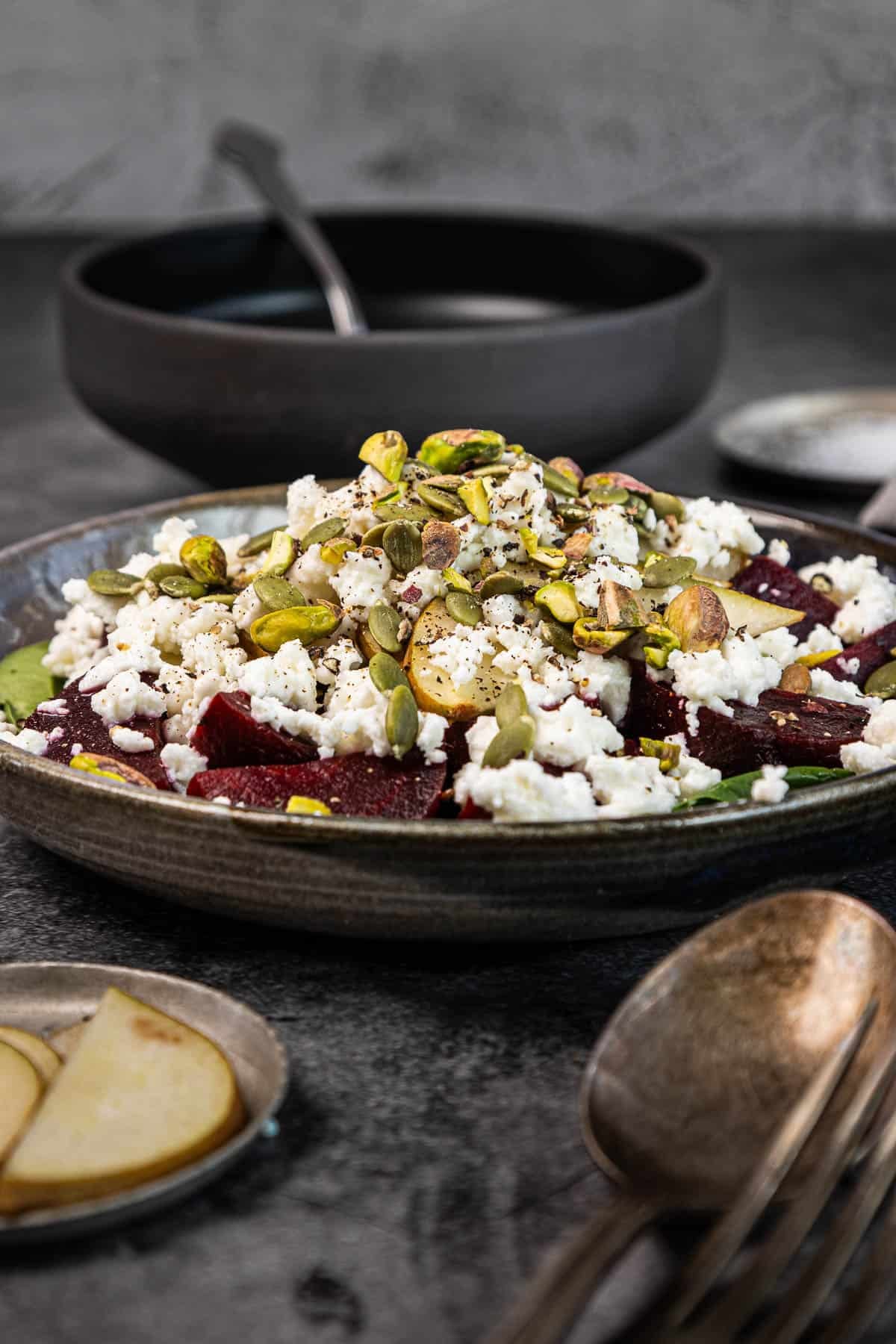 A salad with beet slices, crumbled cheese, and seeds on a gray plate, with cutlery and a small dish nearby.
