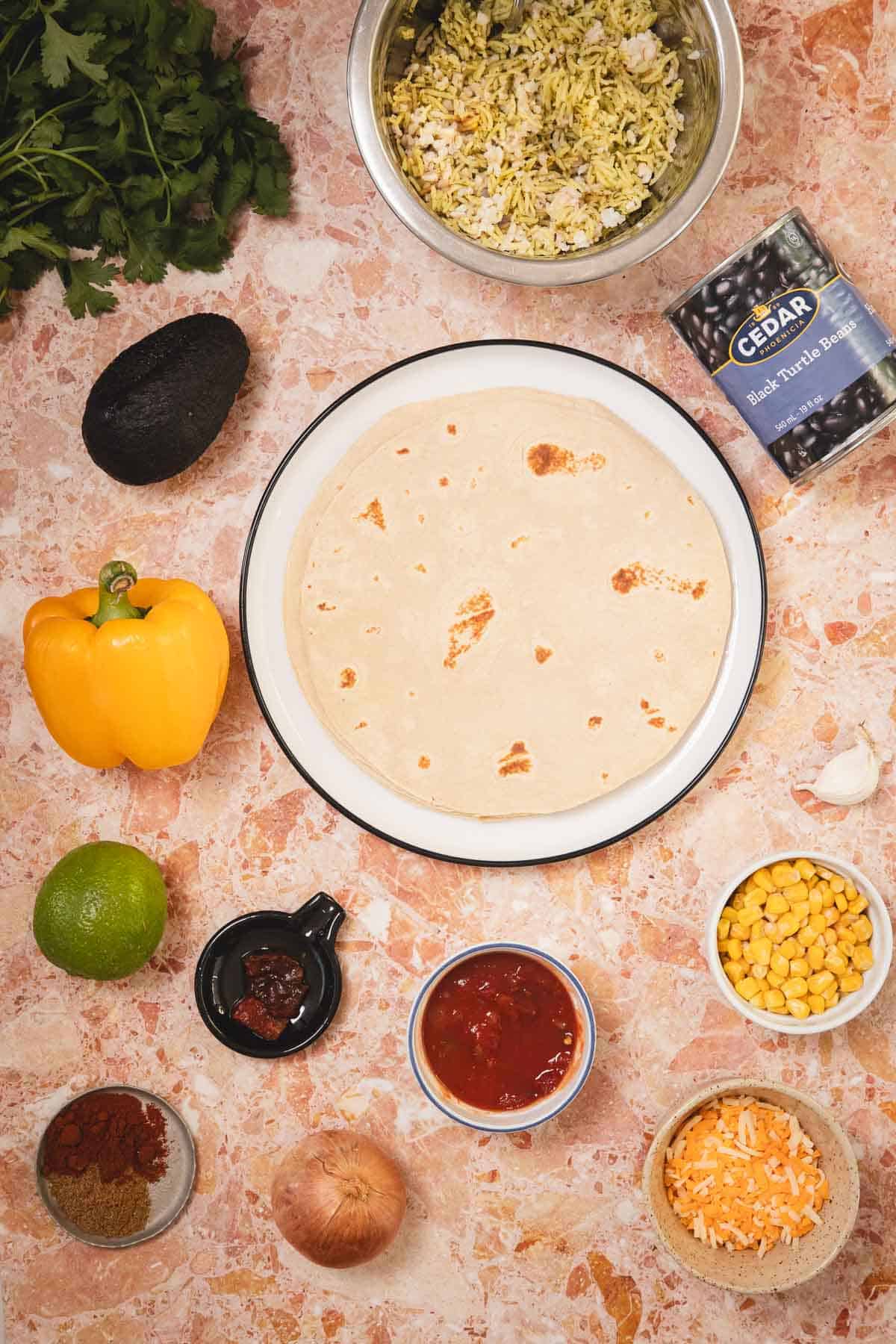 Flat lay of burrito ingredients: tortilla, rice, beans, avocado, yellow pepper, lime, corn, salsa, onion, and spices.