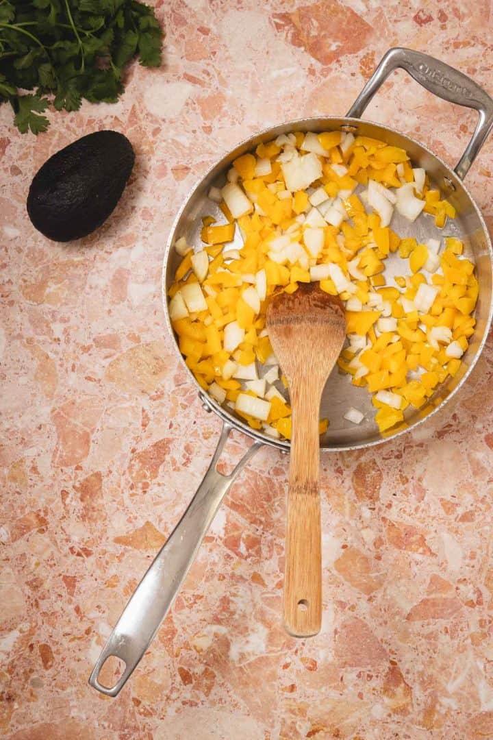 A pan with chopped yellow bell peppers and onions, a wooden spoon, avocado, and herbs on a marble counter.