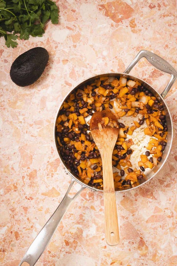 A skillet with sautéed onions, black beans, and sweet potatoes on a marble countertop; avocado nearby.