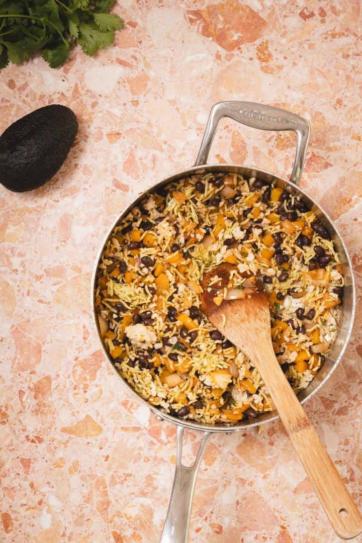 A pot of rice with black beans and vegetables, mixed with a wooden spoon, on a marble countertop.