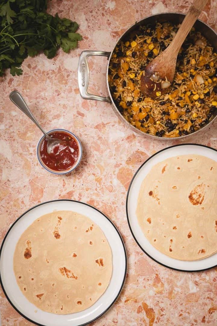 Two plates with tortillas, a bowl of salsa, and a pan of rice, corn, and veggies on a pink countertop.