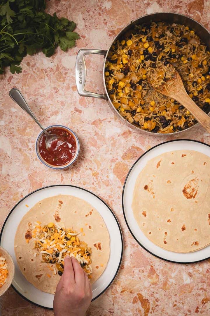 A hand fills a tortilla with a mixture of beans, corn, and cheese; sauce and a skillet are nearby.
