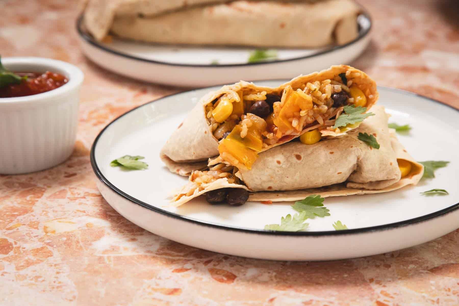 A burrito cut in half, filled with rice, beans, corn, and peppers, on a plate with cilantro garnish.