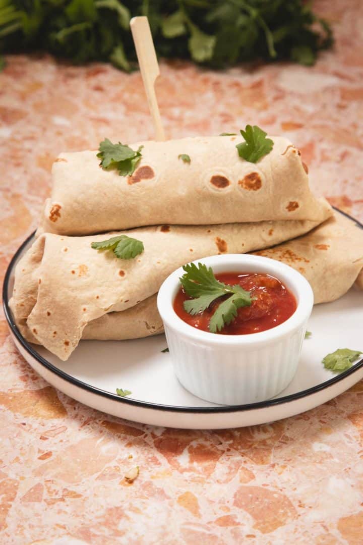 Two burritos on a plate with fresh cilantro, served with a small dish of salsa on a marble table.