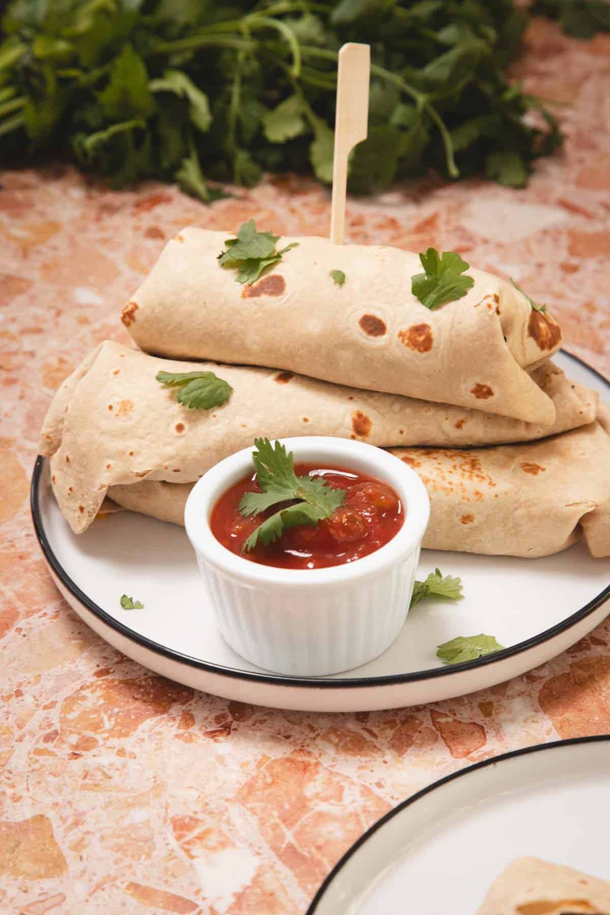 Three rolled tortillas garnished with cilantro on a plate, served with a small bowl of red salsa.