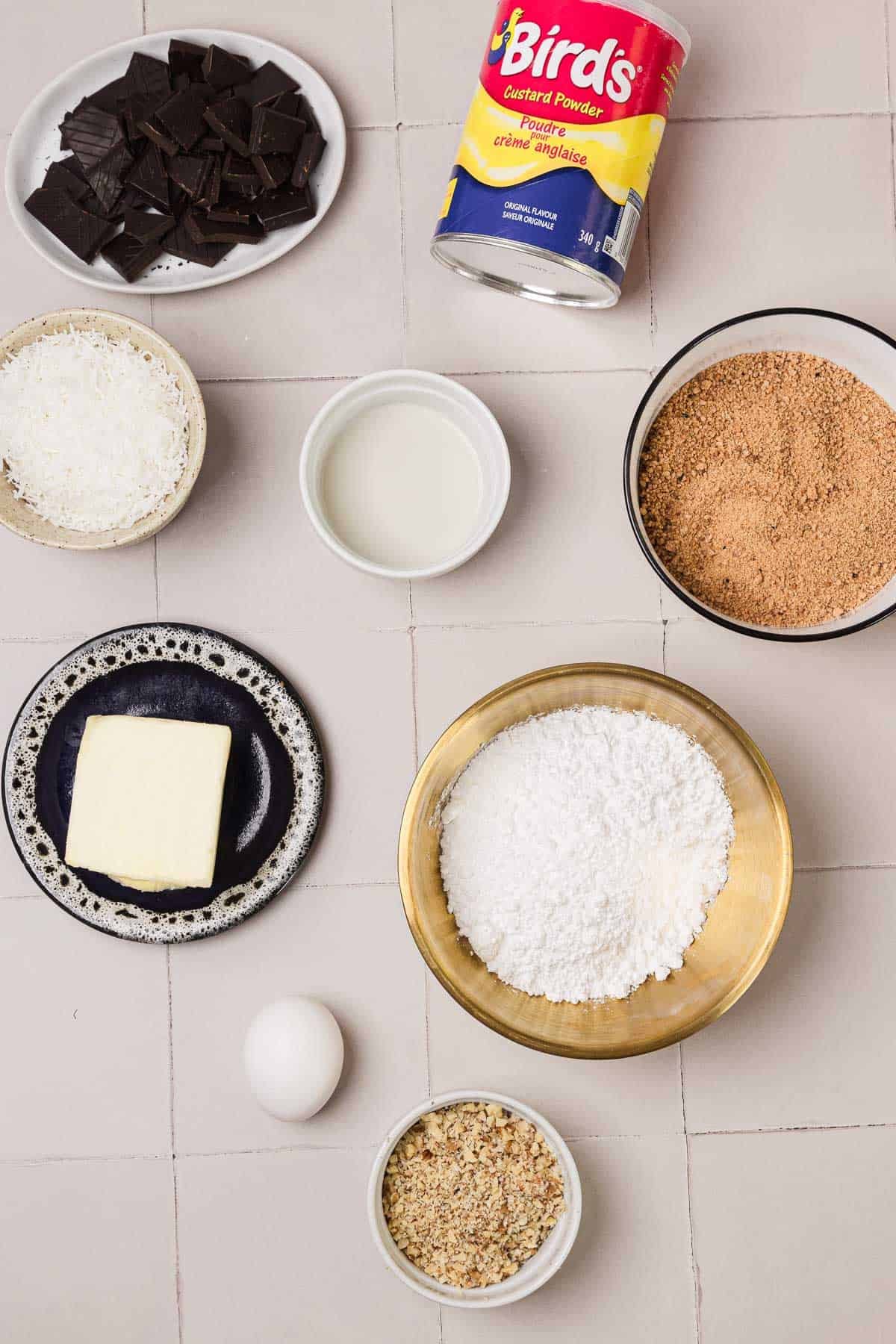 Baking ingredients on a tiled surface: chocolate, custard powder, coconut, butter, egg, milk, nuts, sugar, crumbs.
