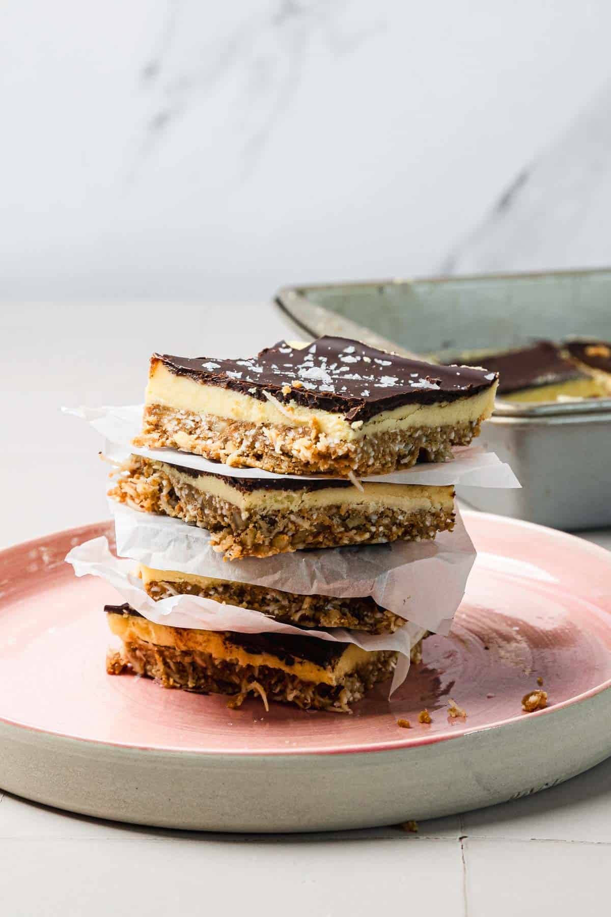 A stack of chocolate caramel bars separated by parchment paper on a pink plate, with more bars in a pan behind.