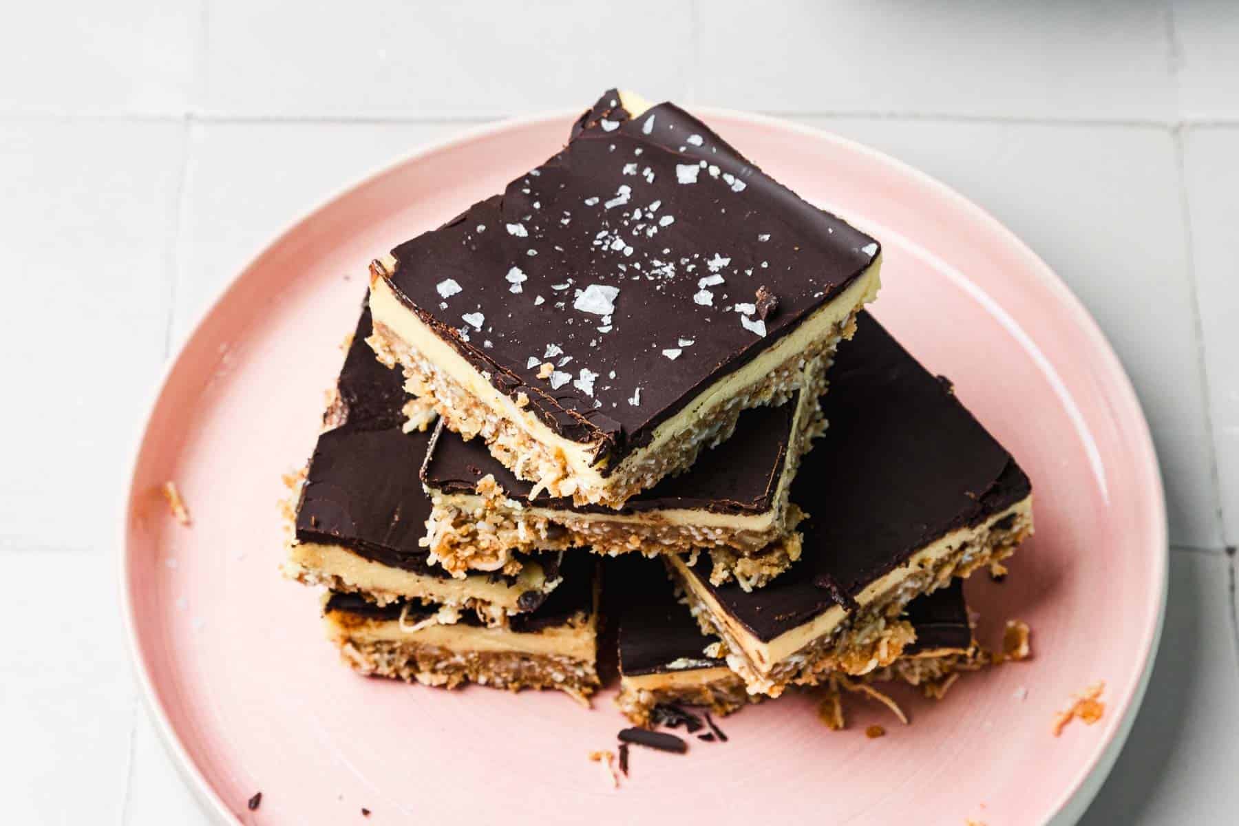Stack of chocolate-topped dessert bars with a crumbly base on a pink plate, sprinkled with sea salt flakes.