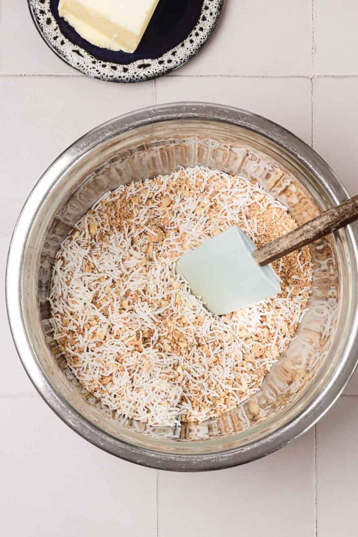 A metal bowl with shredded coconut and brown sugar mixture, and a spatula; a plate of butter is nearby.