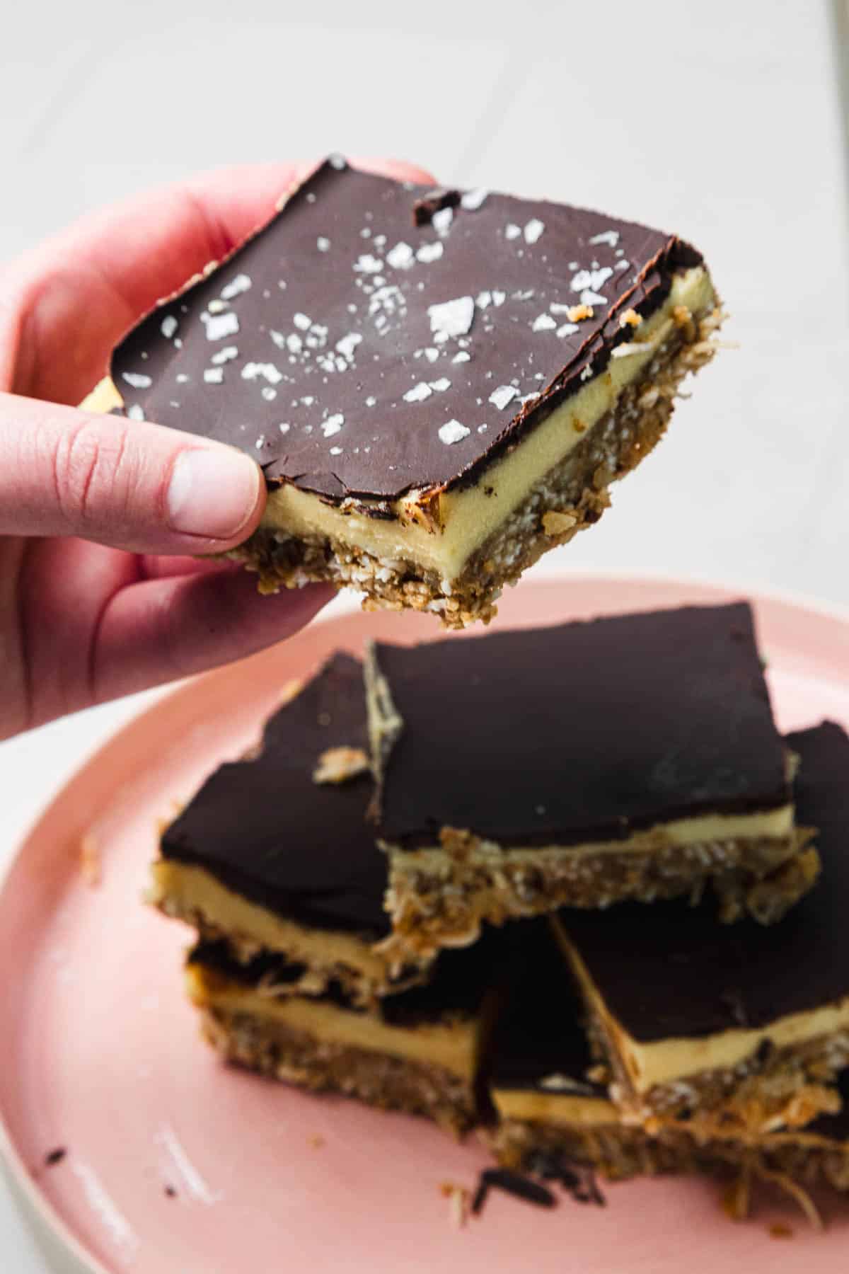 A hand holds a chocolate-topped dessert bar above a pink plate stacked with more bars.