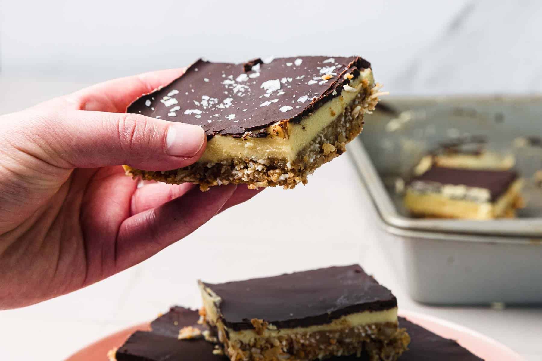 A hand holds a layered dessert bar with a chocolate top; more bars are on a plate and in a baking pan.