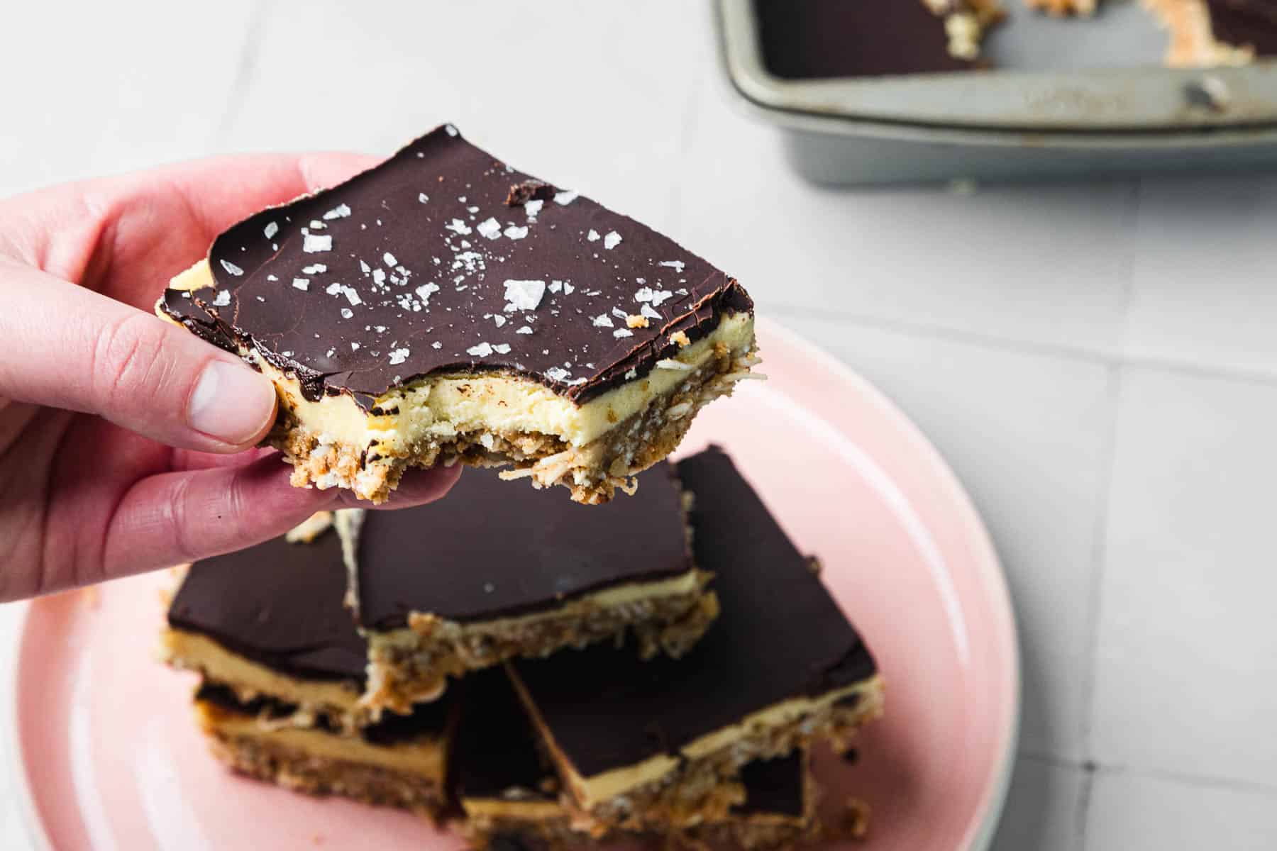 A hand holds a chocolate-topped dessert bar over a plate with more bars and a baking pan in the background.