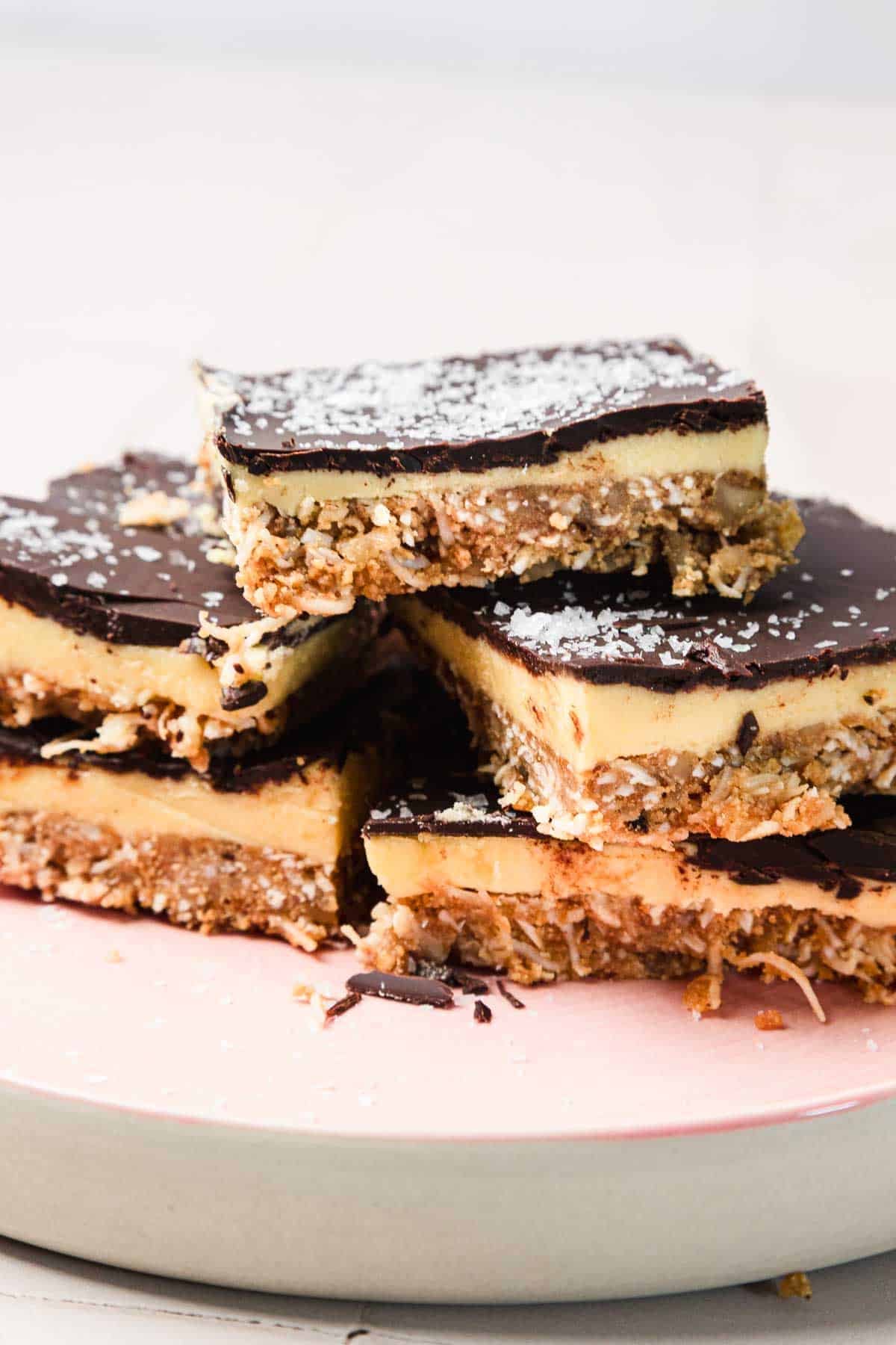 Four stacked Nanaimo bars with chocolate topping and coconut, on a pink plate.