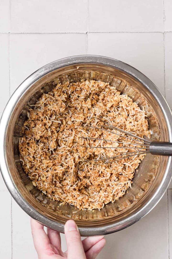 A hand holding a metal bowl of shredded, seasoned mixture being whisked, on a tiled surface.