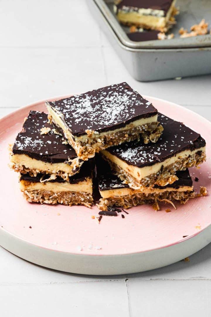 Four chocolate-topped dessert bars stacked on a pink plate, with coconut sprinkled on top.