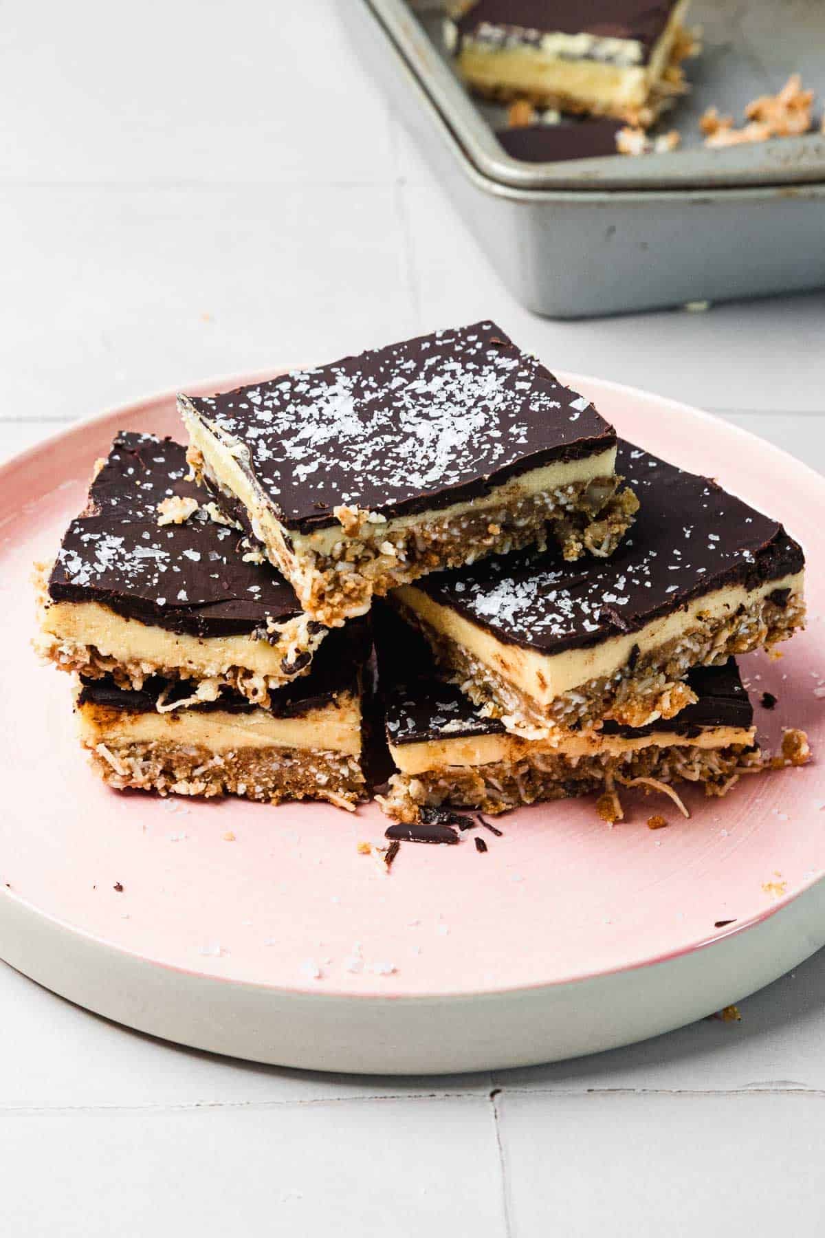 Four chocolate-topped dessert bars stacked on a pink plate, with coconut sprinkled on top.