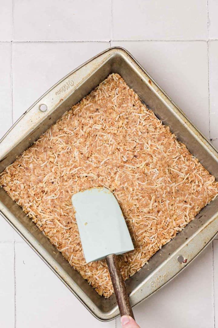 A hand uses a spatula to press coconut crust into a square metal baking pan on a tiled surface.