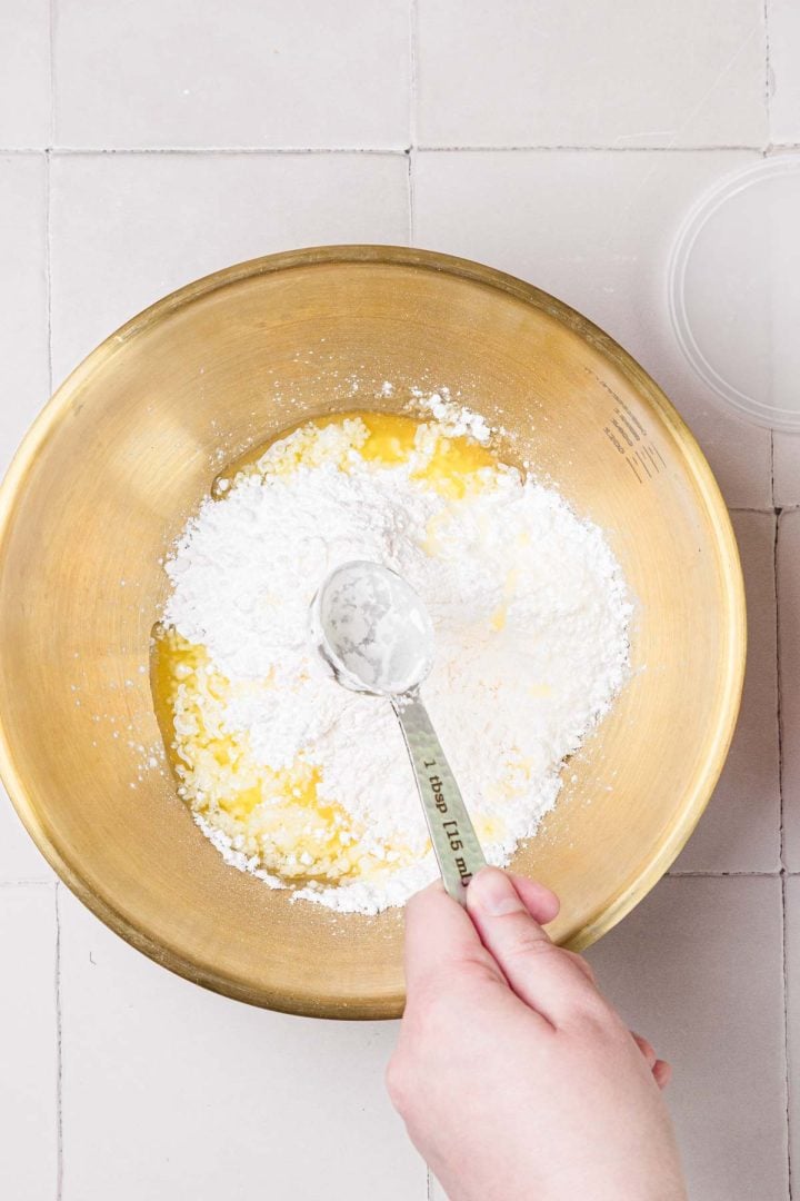 A hand holds a measuring spoon over a mixing bowl with flour, sugar, and melted butter on a tiled surface.