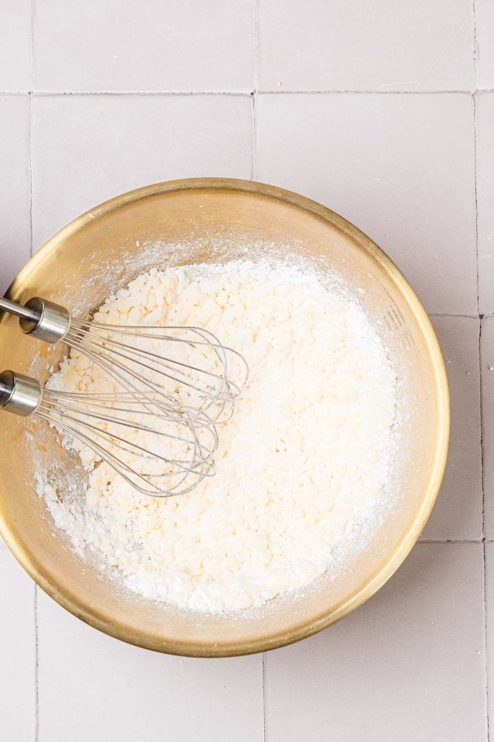 A metal bowl with flour mixture and two whisks on a light tiled surface, viewed from above.