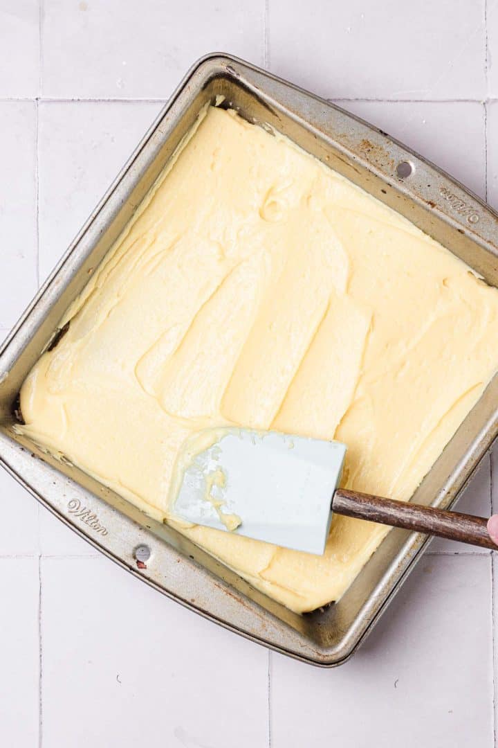 A spatula spreads yellow cake batter evenly in a square metal baking pan on a tiled surface.