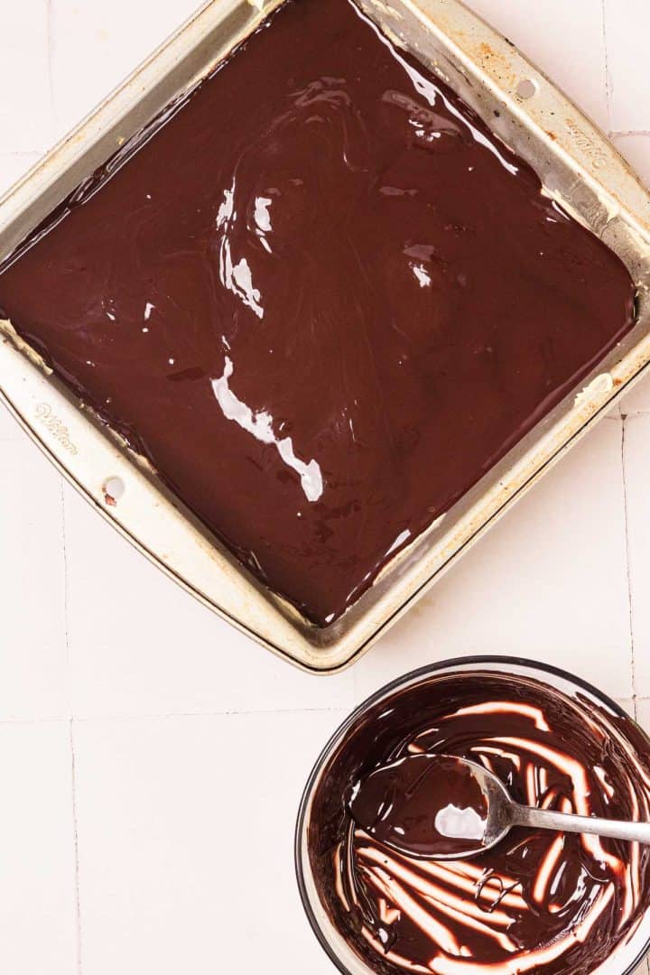 A pan of melted chocolate next to a bowl of chocolate with a spoon on a white surface.