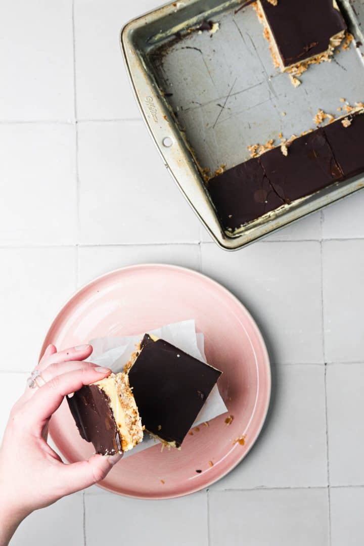 A hand holds a chocolate-topped dessert bar above a pink plate, with more bars in a baking pan nearby.