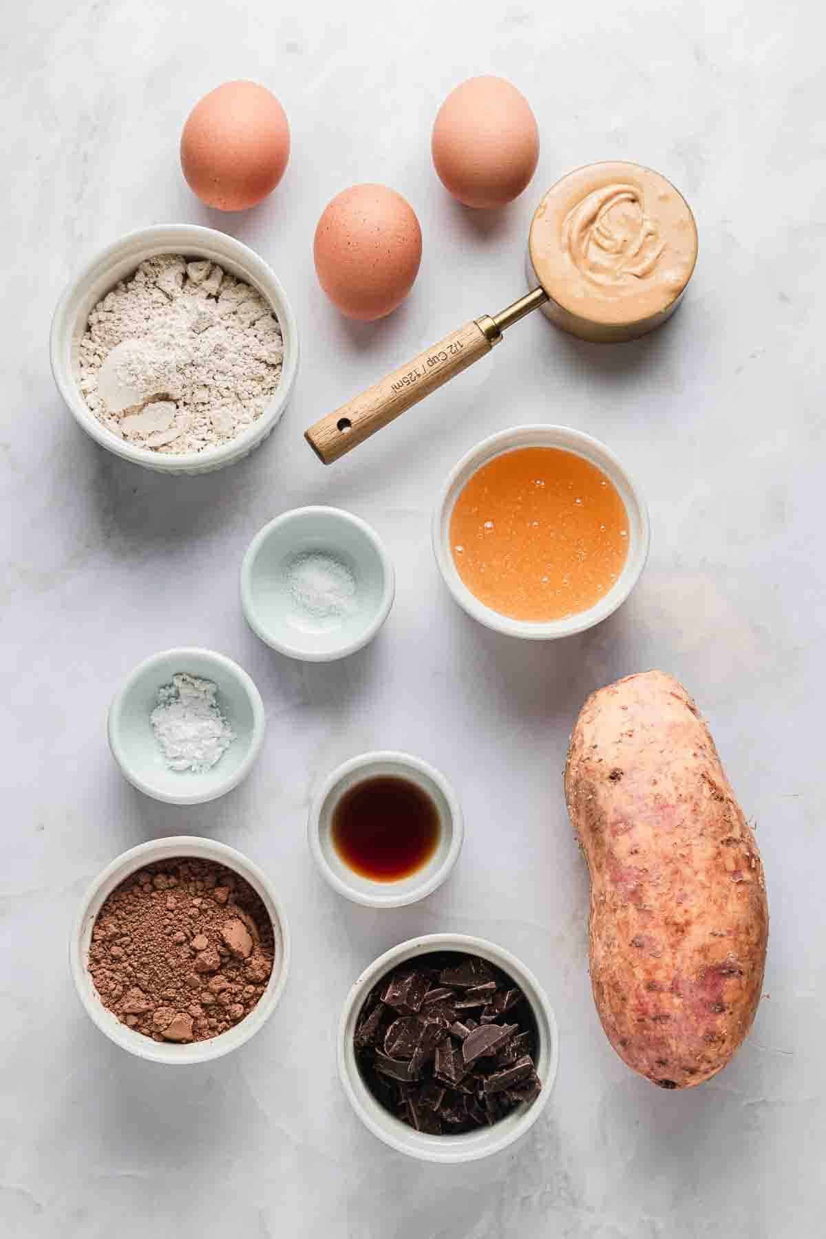 Baking ingredients on a white surface: eggs, flour, sweet potato, cocoa, chocolate, syrup, peanut butter, and vanilla.