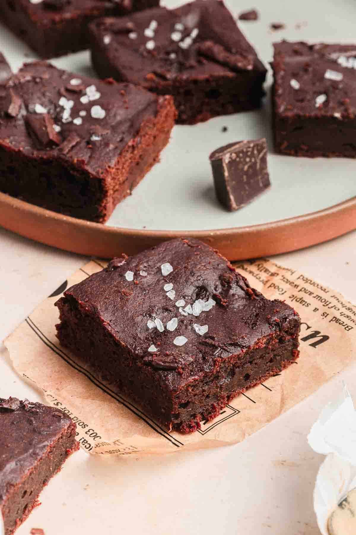 A thick, fudgy chocolate brownie topped with sea salt flakes on parchment paper, with more brownies on a plate.