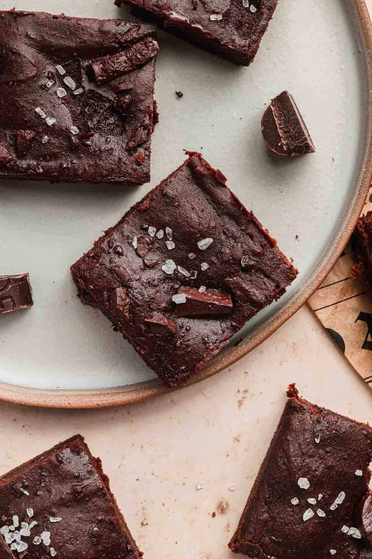Brownies topped with sea salt flakes and chocolate chunks on a round plate, with some pieces on the table.