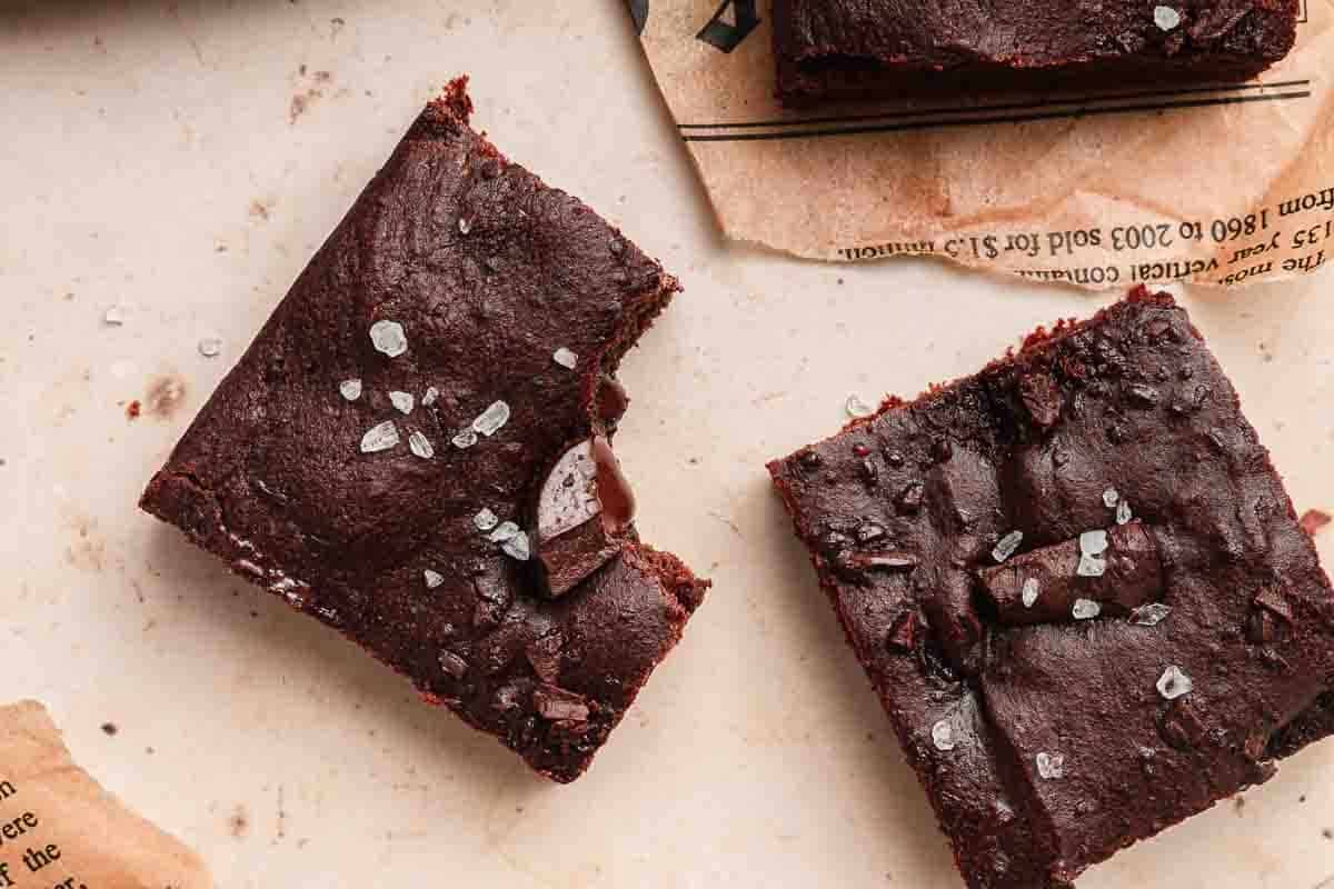 Two fudgy brownies topped with sea salt, one with a bite taken out, on parchment and a light surface.