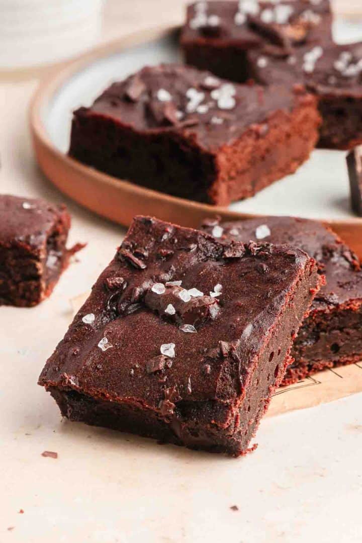 Chocolate brownies topped with flaky sea salt, stacked on a light surface with more brownies on a tray in the background.