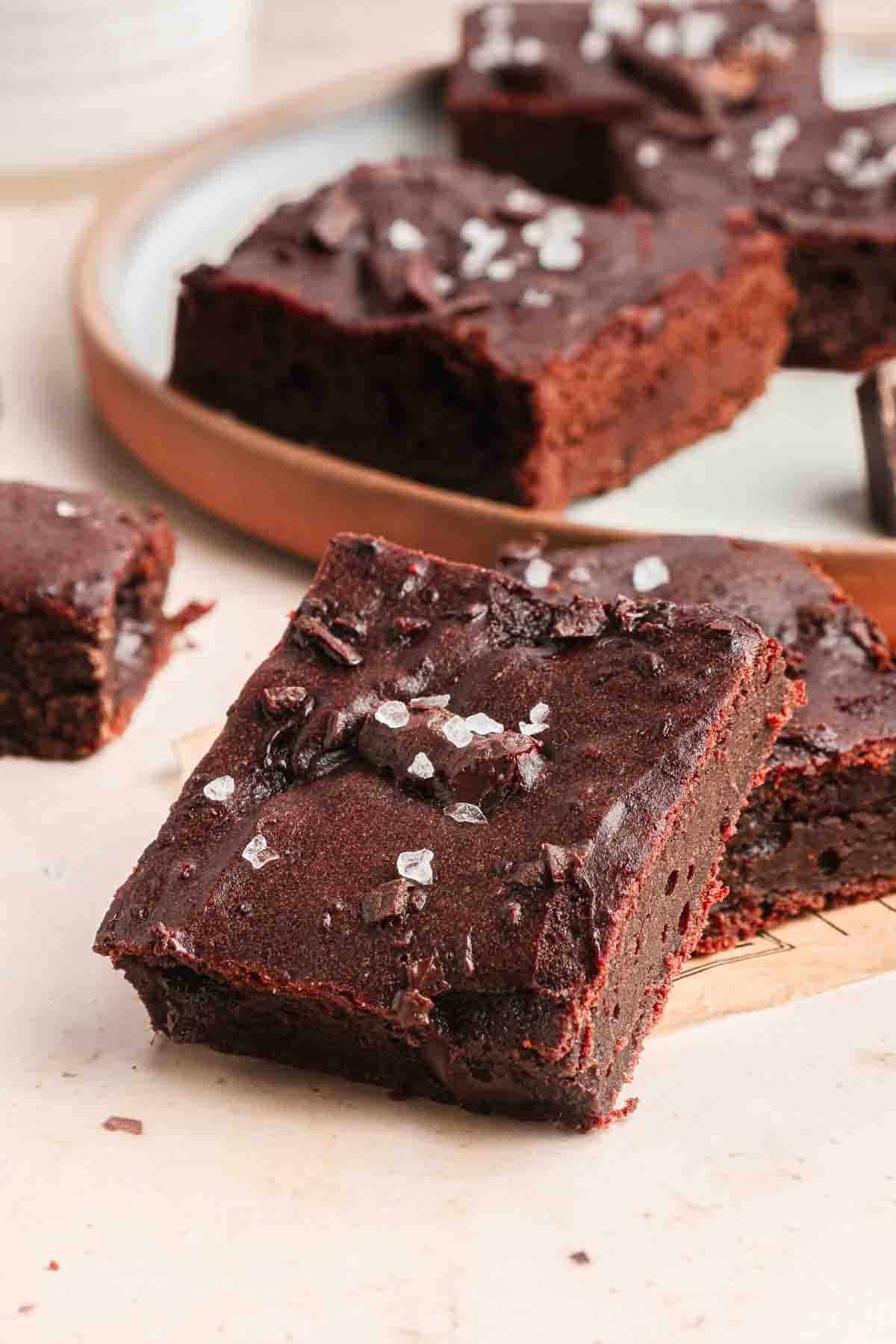 Chocolate brownies topped with flaky sea salt, stacked on a light surface with more brownies on a tray in the background.