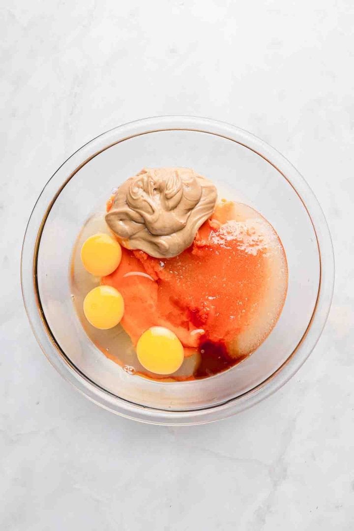 A glass bowl with eggs, brown sugar, vanilla, pumpkin puree, and a dollop of nut butter on a white surface.