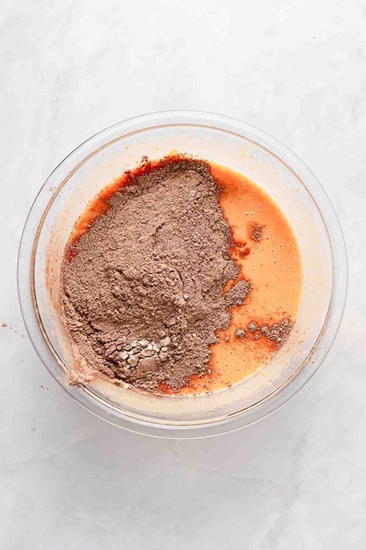 A glass bowl with cocoa powder and liquid ingredients being mixed for baking on a light surface.