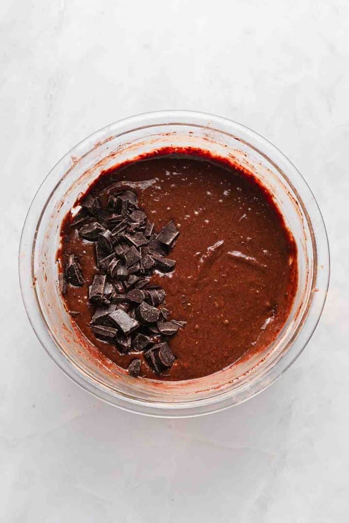 Glass bowl with chocolate brownie batter and chopped dark chocolate on a white marble surface.