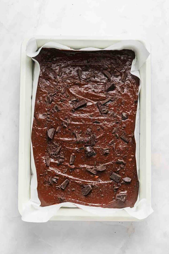 Unbaked brownie batter with chocolate chunks in a parchment-lined baking pan on a light surface.