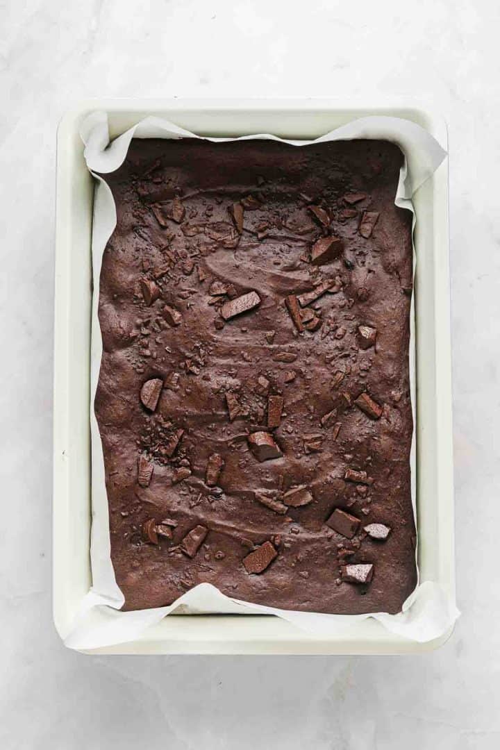 Unbaked brownie batter in a parchment-lined square baking pan with chocolate chunks on top.