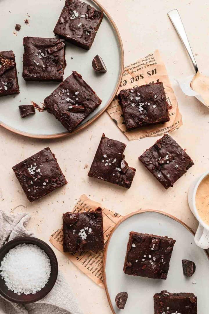 Plates of chocolate brownies topped with sea salt, with coffee and a bowl of sea salt on a light surface.